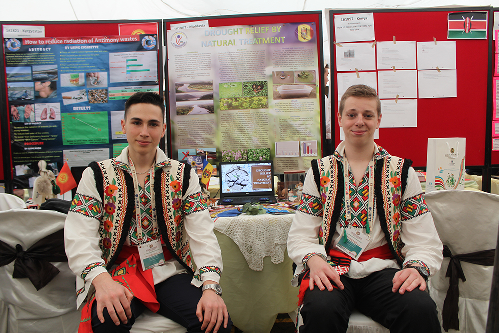 Two students in traditional dress seated at their project stand